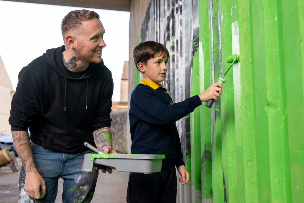 Child in school uniform painting a corrugated wall bright green with a roller, assisted by an artist holding a paint tray.