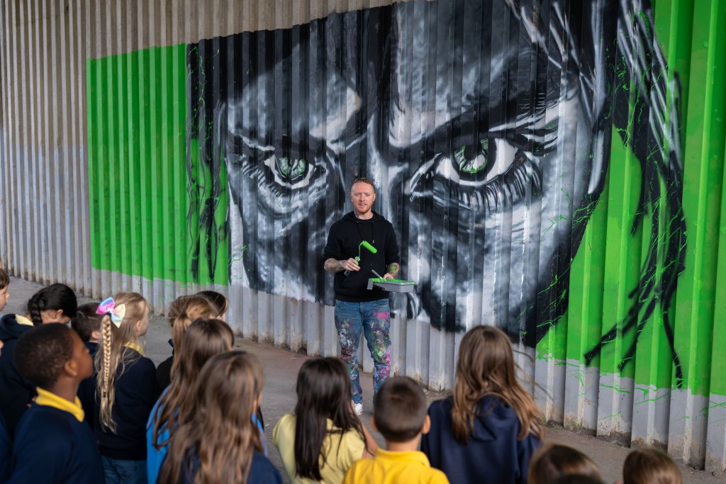 Artist holding a paint roller while explaining a large mural with green eyes to a group of schoolchildren.