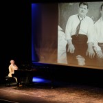 Neil Brand performing on stage with a large black‑and‑white Laurel and Hardy image projected on the screen behind him.