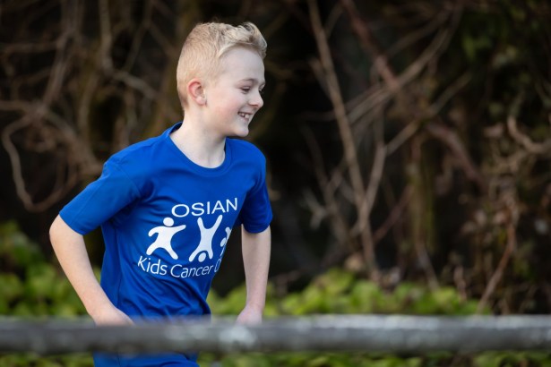 Close‑up of Osian running in a park wearing a blue Kids Cancer Charity T‑shirt.
