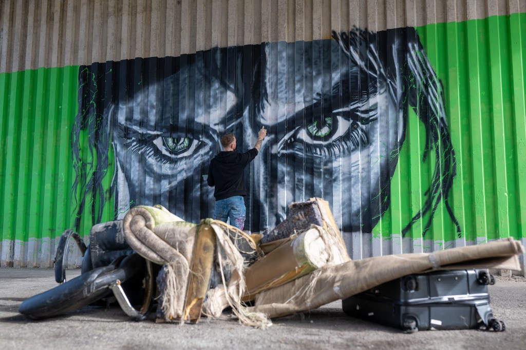 Artist spray‑painting a large mural with green eyes while discarded furniture and waste sit in the foreground.