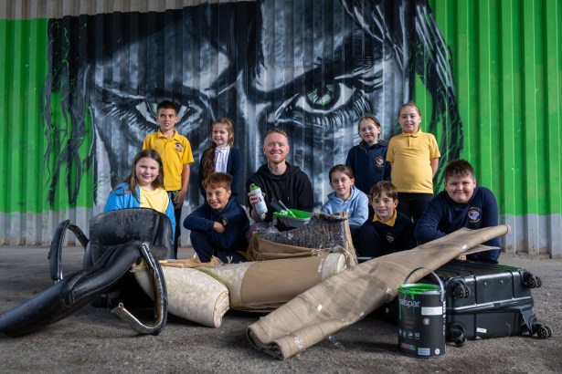 Group of schoolchildren and an artist posing in front of a mural with green eyes, with dumped items arranged in front.