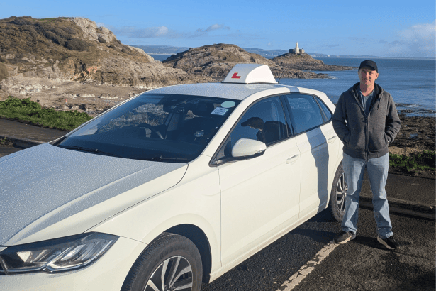 Driving instructor Mike Colwill standing beside his branded training car near Mumbles Lighthouse.