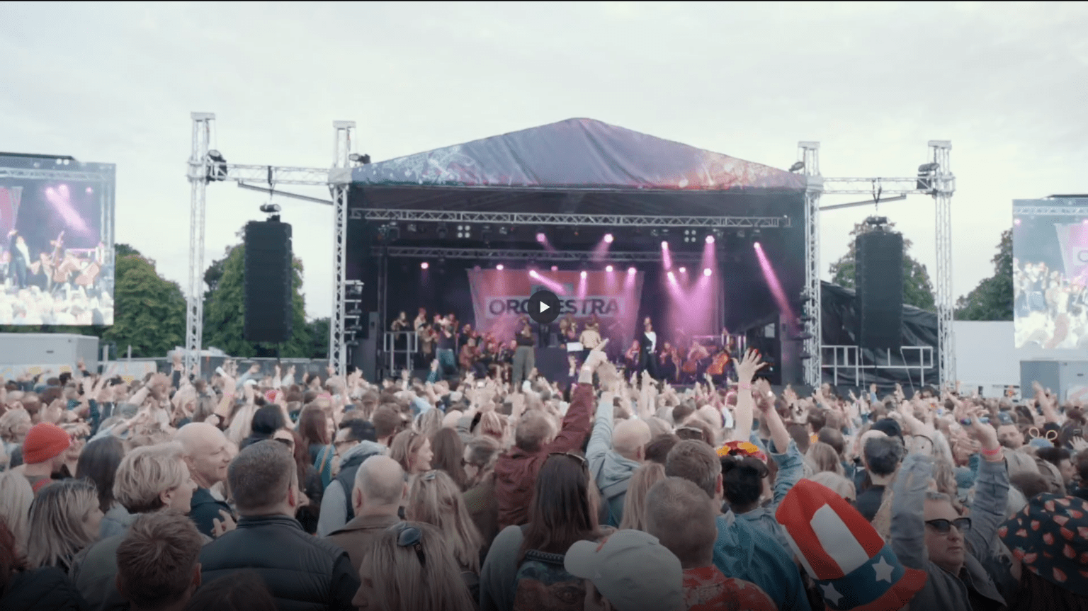 Wide-angle photo of The Ibiza Orchestra performing live at a packed outdoor concert, with large screens and cheering crowds.