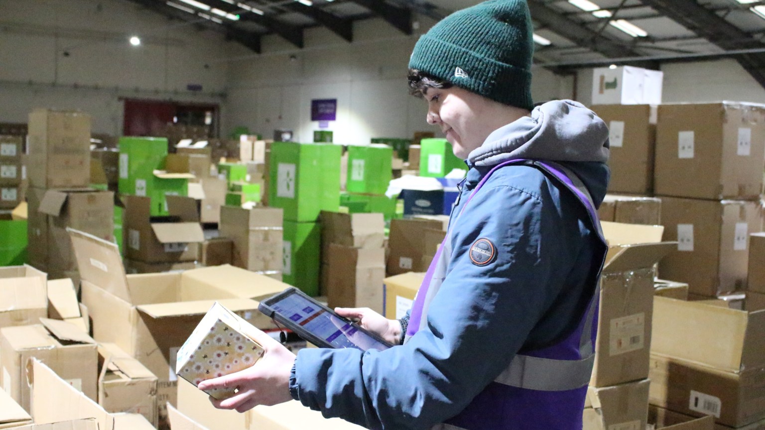 Warehouse worker in a blue coat, purple hi‑vis and green beanie holding a tablet and a boxed item among large cardboard boxes at Something Different Wholesale.