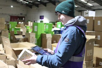 Warehouse worker in a blue coat, purple hi‑vis and green beanie holding a tablet and a boxed item among large cardboard boxes at Something Different Wholesale.