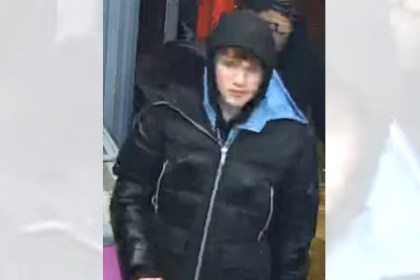 CCTV image of a male wearing a black puffer jacket with hood up, standing indoors near a doorway.