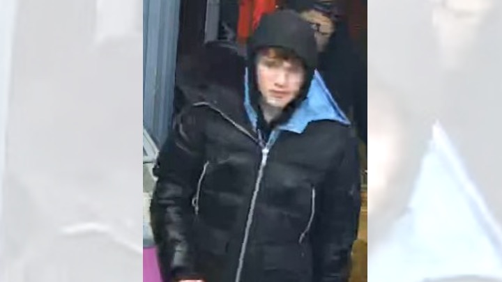 CCTV image of a male wearing a black puffer jacket with hood up, standing indoors near a doorway.