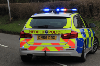Rear view of a Dyfed‑Powys Police car with bilingual markings and blue emergency lights, parked on the roadside.
