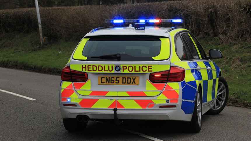 Rear view of a Dyfed‑Powys Police car with bilingual markings and blue emergency lights, parked on the roadside.
