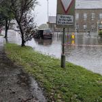 Surface water flooding across the Killay Square junction, with cars driving slowly through deep pooled water.