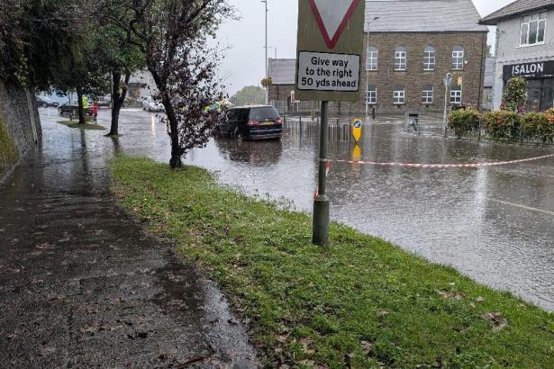Surface water flooding across the Killay Square junction, with cars driving slowly through deep pooled water.
