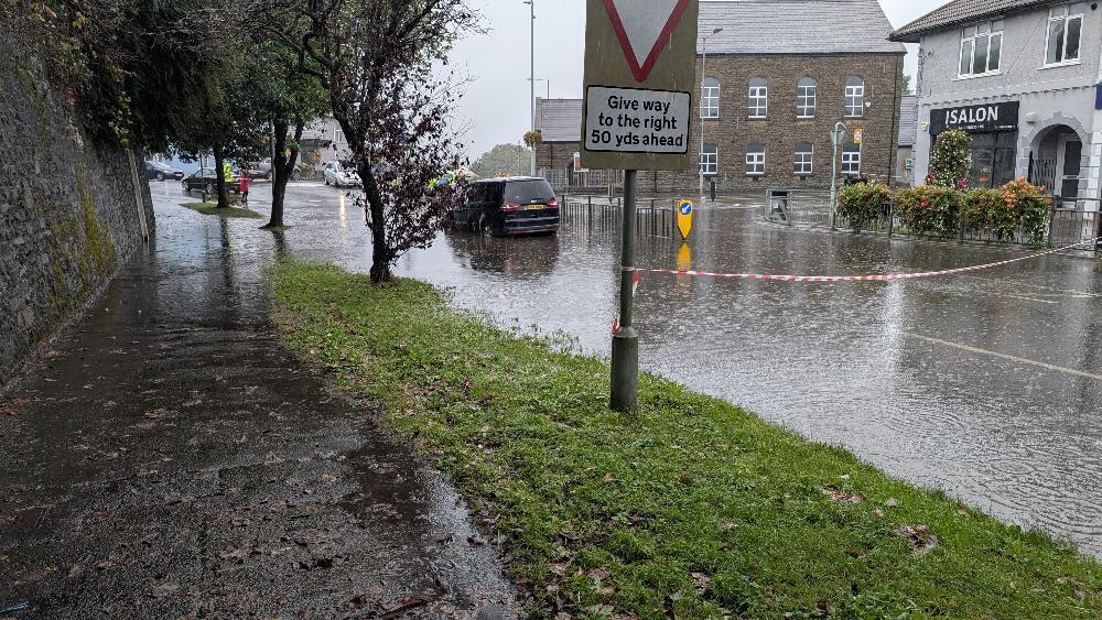 Surface water flooding across the Killay Square junction, with cars driving slowly through deep pooled water.