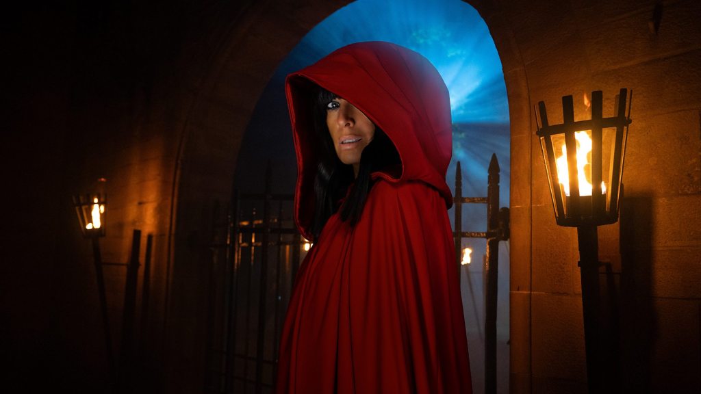 Claudia Winkleman in the signature red hood from The Traitors, standing in a dramatic castle setting.