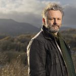 Michael Sheen standing outdoors with a backdrop of rolling green hills in South Wales.