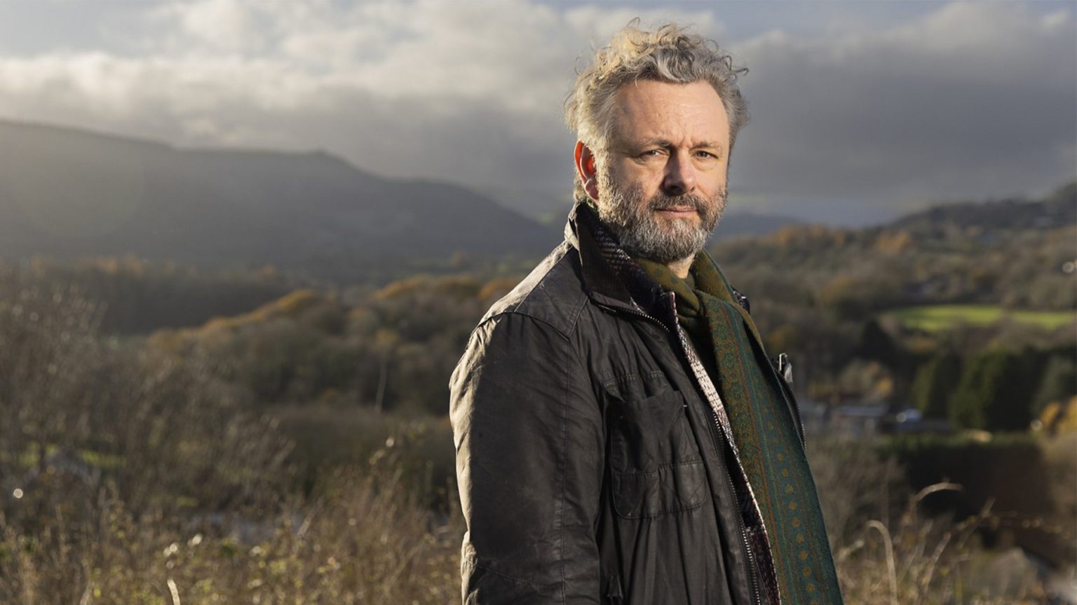 Michael Sheen standing outdoors with a backdrop of rolling green hills in South Wales.
