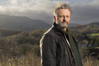 Michael Sheen standing outdoors with a backdrop of rolling green hills in South Wales.