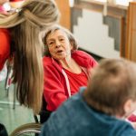 Two older women sitting at a table in a care home, one in a wheelchair, with a staff member speaking to them.