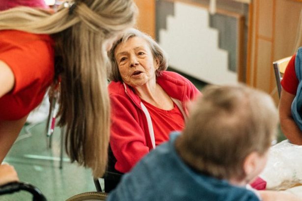 Two older women sitting at a table in a care home, one in a wheelchair, with a staff member speaking to them.