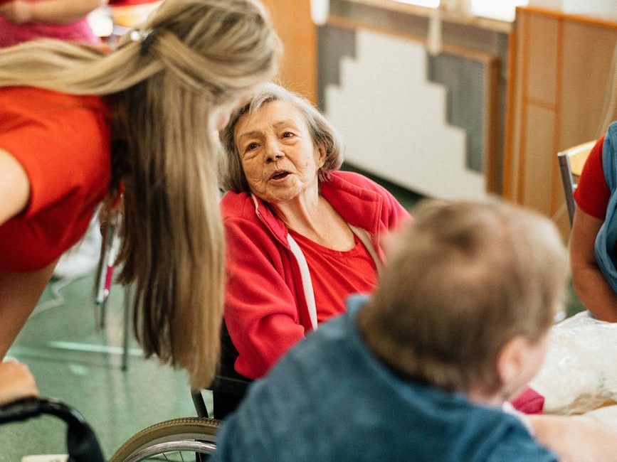 Two older women sitting at a table in a care home, one in a wheelchair, with a staff member speaking to them.