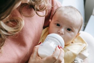 cute newborn drinking milk from bottle in hands of crop mom