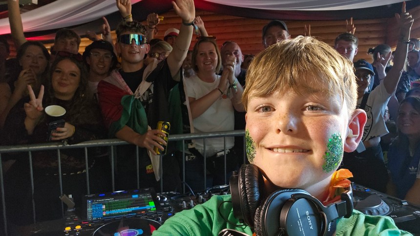 Efan Electro taking a selfie at the DJ desk with glitter on his cheeks, headphones around his neck and a crowd behind him.