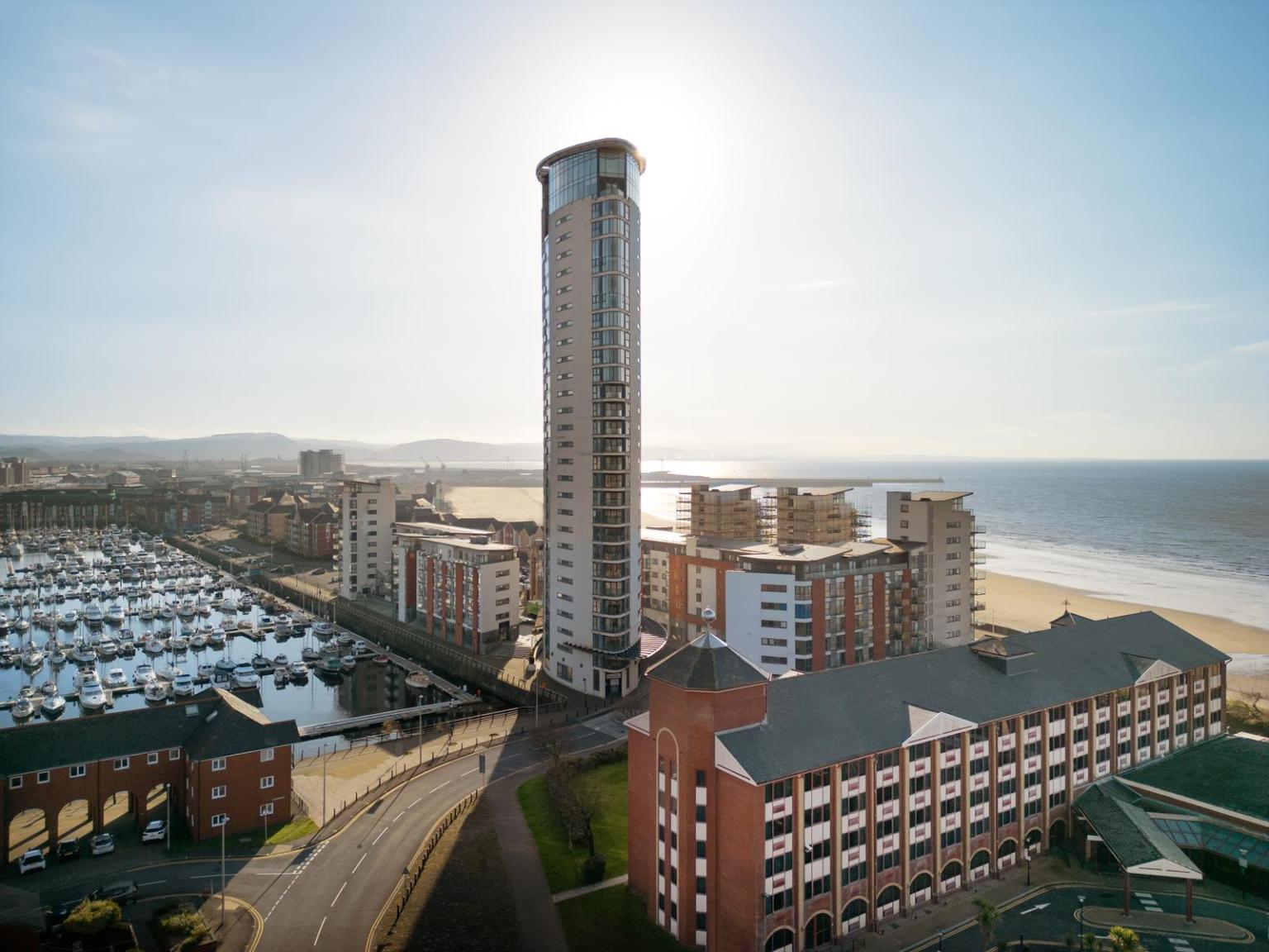Aerial view of the Delta Hotels by Marriott Swansea in the Maritime Quarter, a red brick building in the foreground with the tall Meridian Tower behind it, Swansea Marina to the left and Swansea Bay stretching into the distance