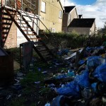 Back lane in Llanelli littered with dumped bags, cans and household waste beneath an external staircase.