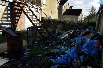 Back lane in Llanelli littered with dumped bags, cans and household waste beneath an external staircase.