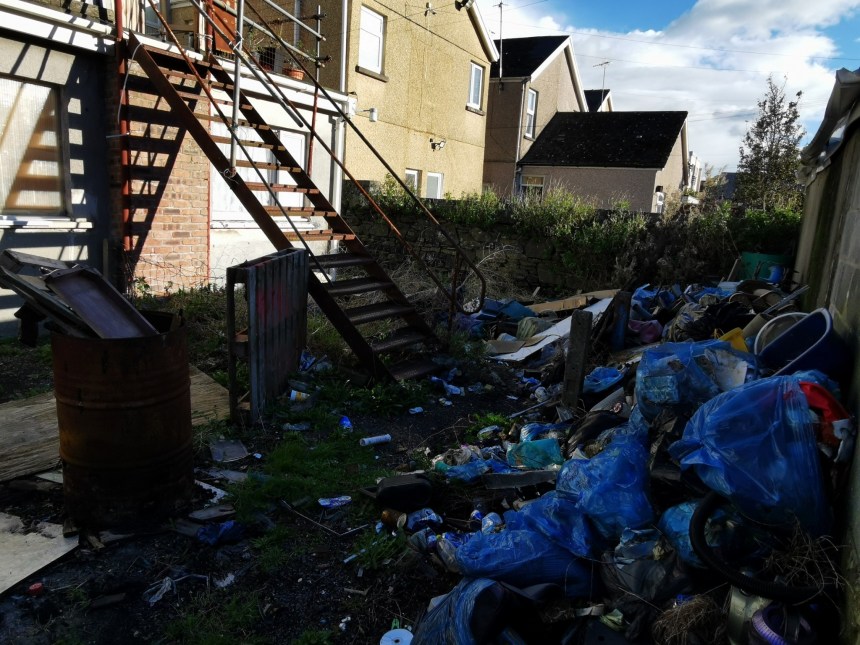 Back lane in Llanelli littered with dumped bags, cans and household waste beneath an external staircase.