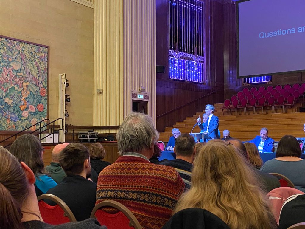 Torsten Bell MP at a podium in Brangwyn Hall addressing a full audience during a public meeting on the future of the Ospreys.