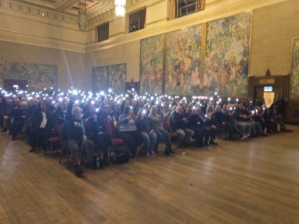 A large audience inside Brangwyn Hall holding up phone lights during a public meeting about the future of the Ospreys.