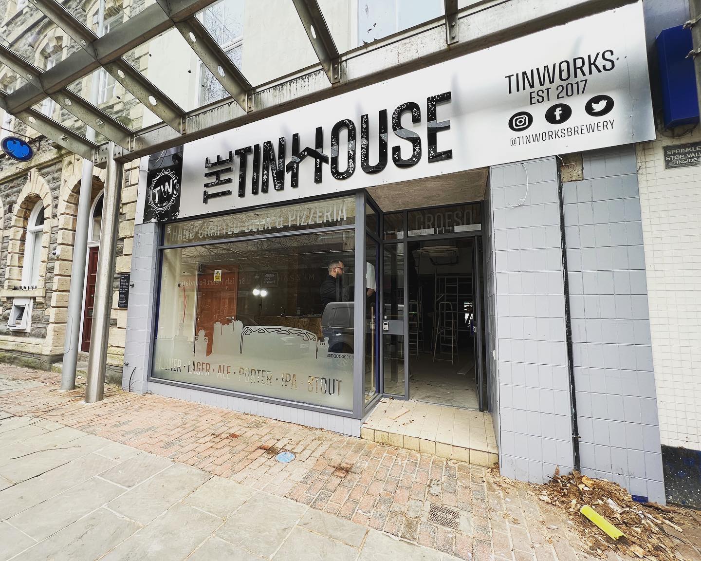 Front view of The Tinhouse taproom in Llanelli, showing the craft beer and pizzeria signage above the entrance and the interior visible through the glass frontage.