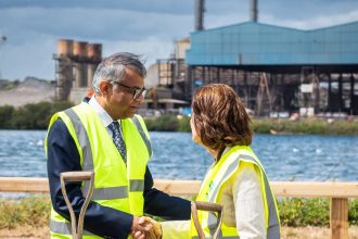 First Minister Eluned Morgan in hi-vis vest shaking hands with Tata Steel UK CEO Rajesh Nair in hi-vis vest at Port Talbot steelworks with industrial buildings visible in background