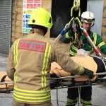 Firefighters and a paramedic use a rope‑based lifting system to move a secured training dummy on a stretcher during a bariatric rescue exercise at a South Wales Fire and Rescue Service facility.