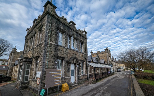 Stone-built Maesteg Hospital with Gothic features, showing main entrance and outpatient signage under cloudy sky.