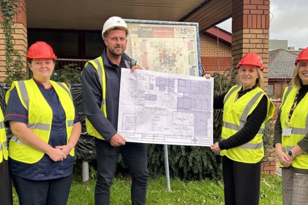 Group of Hywel Dda staff and project leads in hard hats and high‑visibility vests holding refurbishment plans outside the Priory Day Unit at Glangwili Hospital.