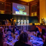 Wide view of Brangwyn Hall during the Swansea Bay Business Awards 2026, with guests seated at candlelit tables and event branding on the main screen.