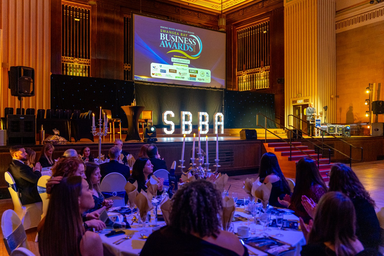 Wide view of Brangwyn Hall during the Swansea Bay Business Awards 2026, with guests seated at candlelit tables and event branding on the main screen.
