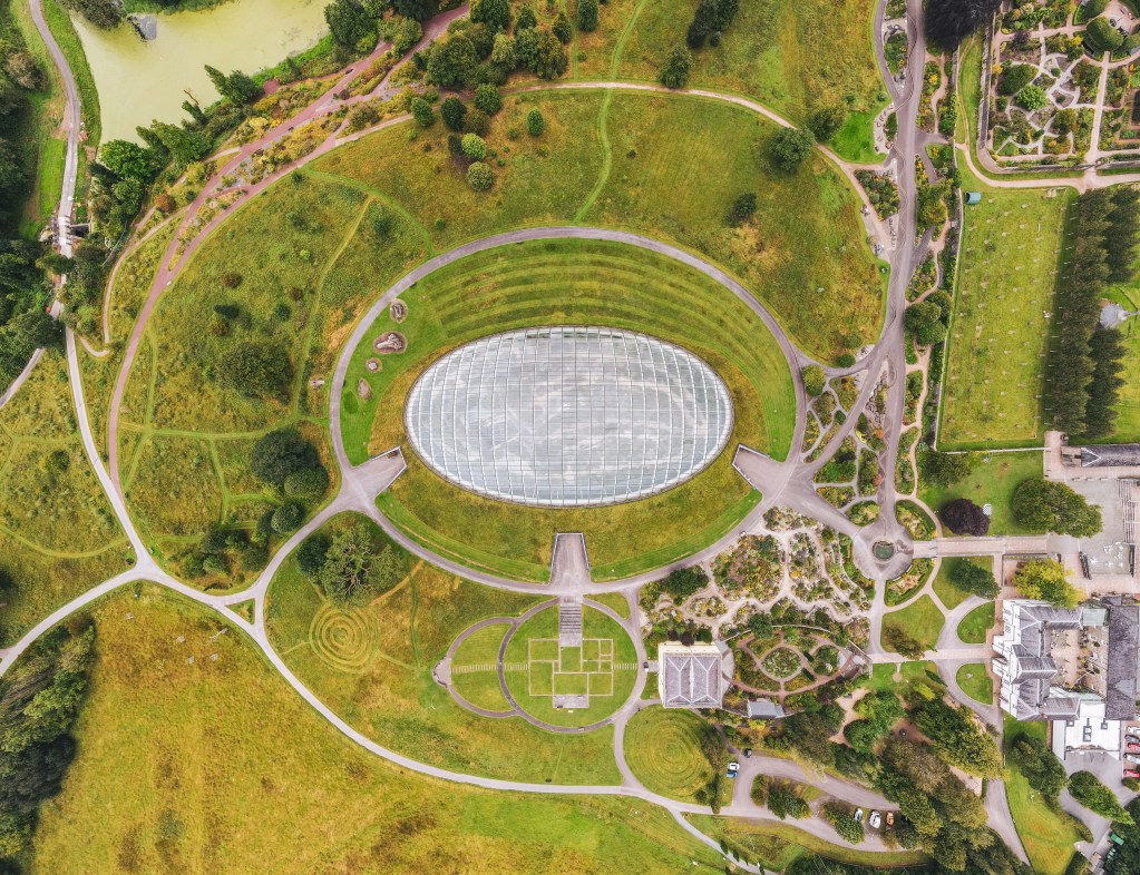 High‑level aerial shot showing the Great Glasshouse, garden structures, lawns and the wider landscape of the National Botanic Garden of Wales.