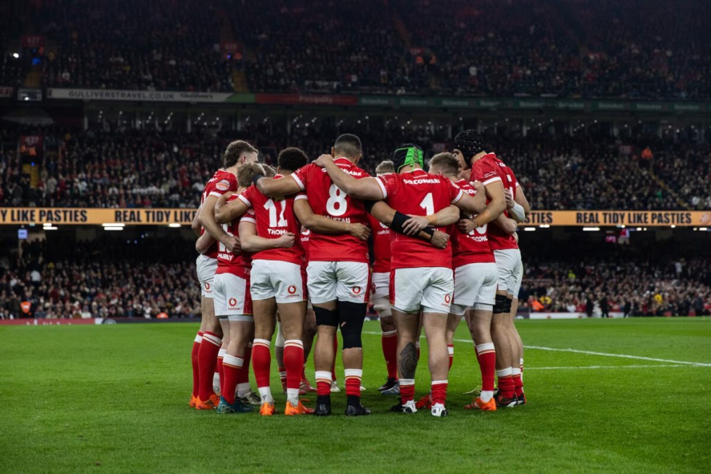 Wales rugby players in a pre‑match huddle on the field with a packed stadium behind them.
