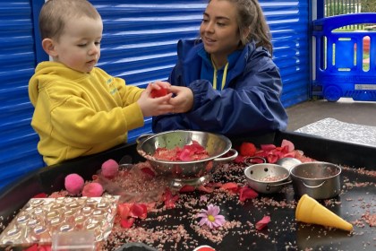 Child and adult interacting at a sensory play table filled with colourful materials at a Flying Start setting.