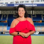 Hayley Parsons MBE standing on the pitch at Cardiff Rugby’s ground in a red dress, with stadium seating and sponsor boards behind her.