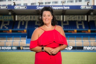 Hayley Parsons MBE standing on the pitch at Cardiff Rugby’s ground in a red dress, with stadium seating and sponsor boards behind her.