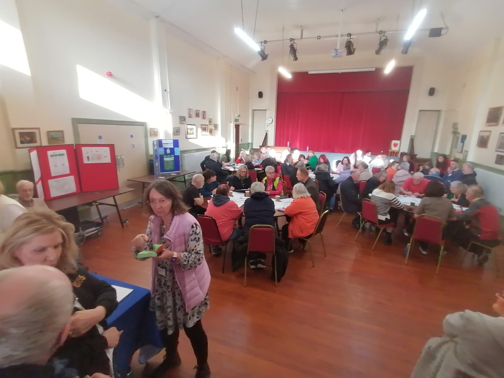 Packed community hall with residents seated at round tables during a Hywel Dda drop-in engagement session about the proposed closure of Meddygfa'r Sarn surgery