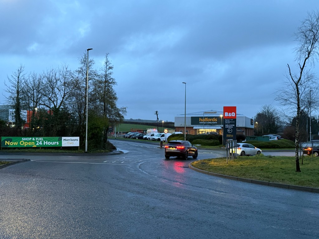 Roundabout at Parc Pensarn with cars passing Halfords, B&Q signage and a Morrisons Daily 24‑hour shop and fuel sign.