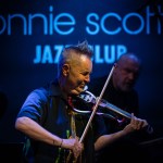 Nigel Kennedy performing on stage at Ronnie Scott’s jazz club, playing electric violin under blue neon lighting.