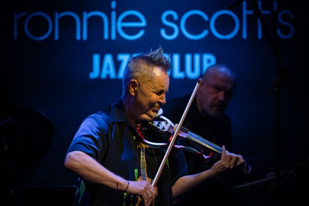 Nigel Kennedy performing on stage at Ronnie Scott’s jazz club, playing electric violin under blue neon lighting.