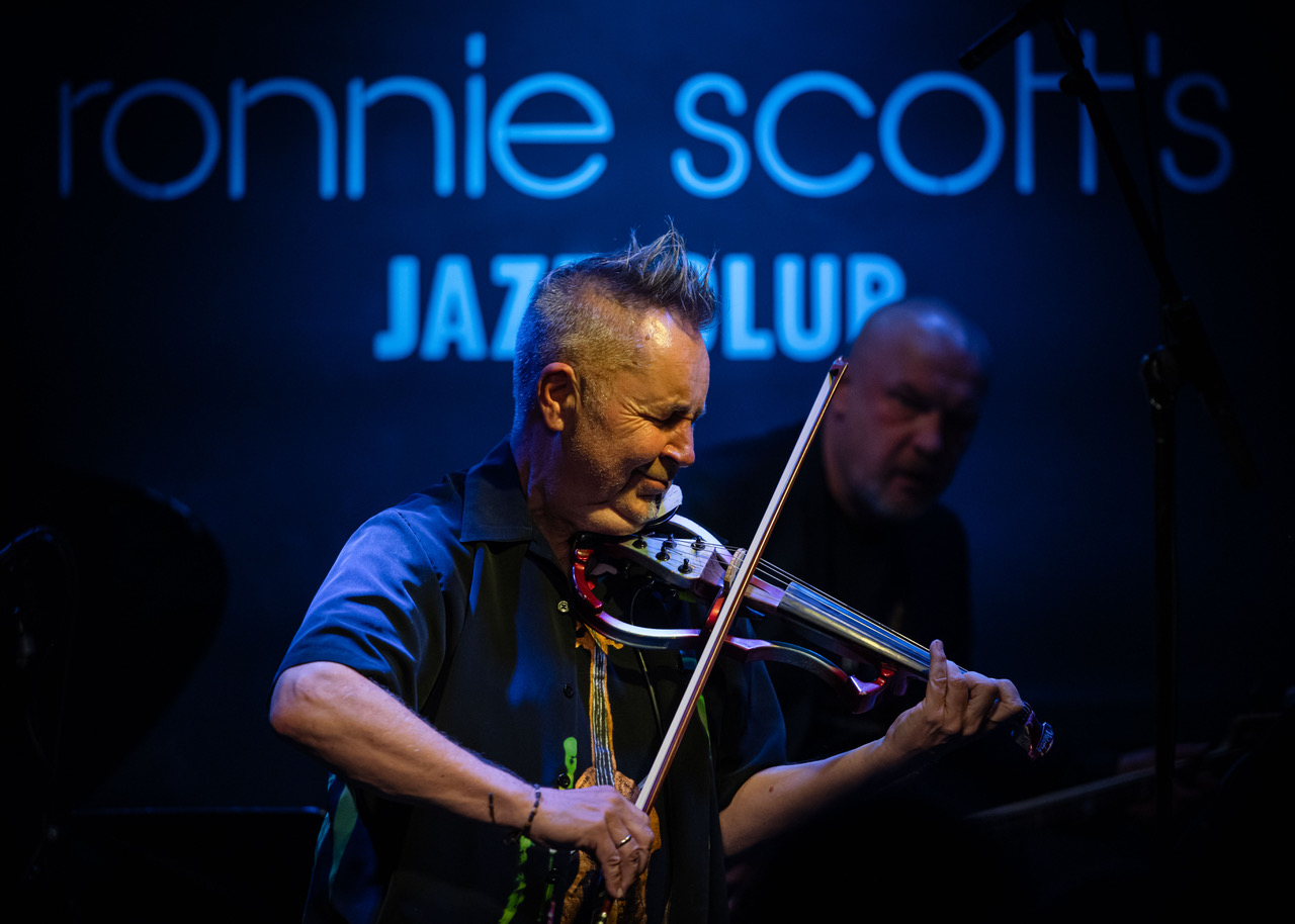 Nigel Kennedy performing on stage at Ronnie Scott’s jazz club, playing electric violin under blue neon lighting.