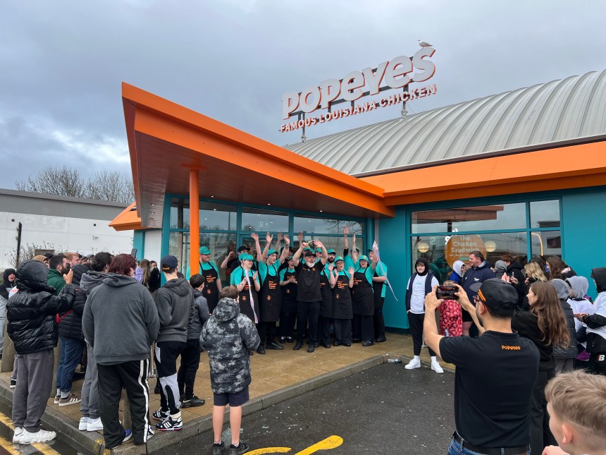 Popeyes staff in teal uniforms celebrating outside the new Llanelli restaurant during the grand opening, with crowds gathered around the entrance.